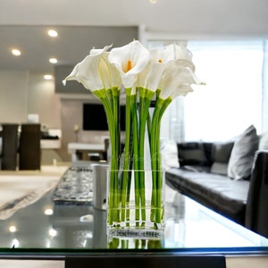 May include: A clear glass vase filled with white calla lilies with green stems. The vase is sitting on a glass table top.