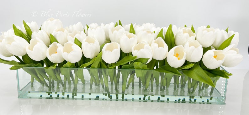 May include: A glass vase filled with white tulips with green leaves. The vase is clear and has a rectangular shape.