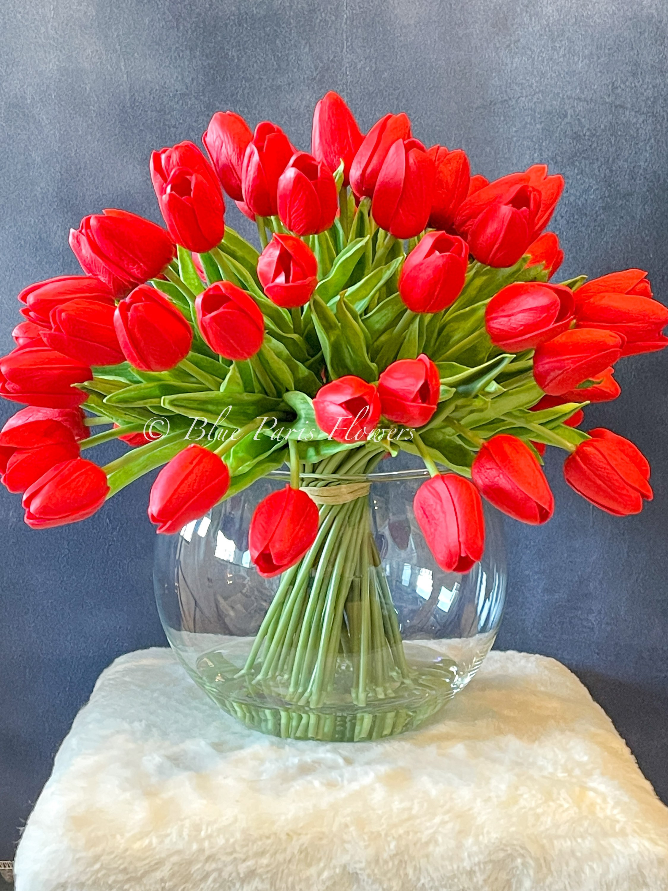 May include: A bouquet of red tulips arranged in a clear glass vase. The vase is sitting on a white fluffy surface.