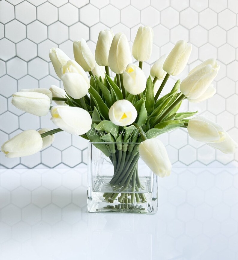 May include: A bouquet of white artificial tulips arranged in a clear glass cube vase. The vase is sitting on a white tiled surface.