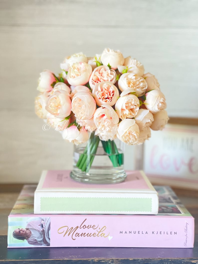 May include: A bouquet of blush pink peonies in a clear glass vase, sitting on top of two pink and green books. The top book has the title "love" and the bottom book has the title "love  Manuela" with the author's name "MANUELA KJEILEN".