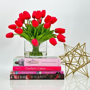 May include: A bouquet of red tulips in a clear glass vase, sitting on a stack of books with titles like "Love Manuela", "Confetti Cakes", "Macarons", and "Herm&eacute;s Macaron". A gold geometric star sculpture is on the right side of the image.