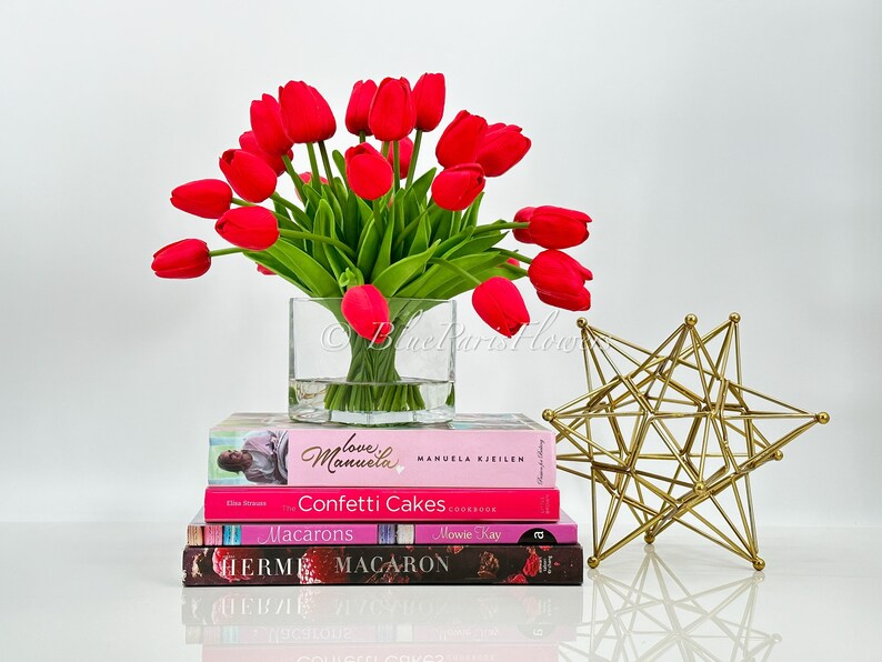 May include: A bouquet of red tulips in a clear glass vase, sitting on a stack of books with titles like "Love Manuela", "Confetti Cakes", "Macarons", and "Herm&eacute;s Macaron". A gold geometric star sculpture is on the right side of the image.