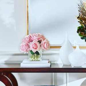 May include: A wooden console table with two shelves, featuring a bouquet of pink roses in a clear glass vase. The table is decorated with books and two white ceramic vases.