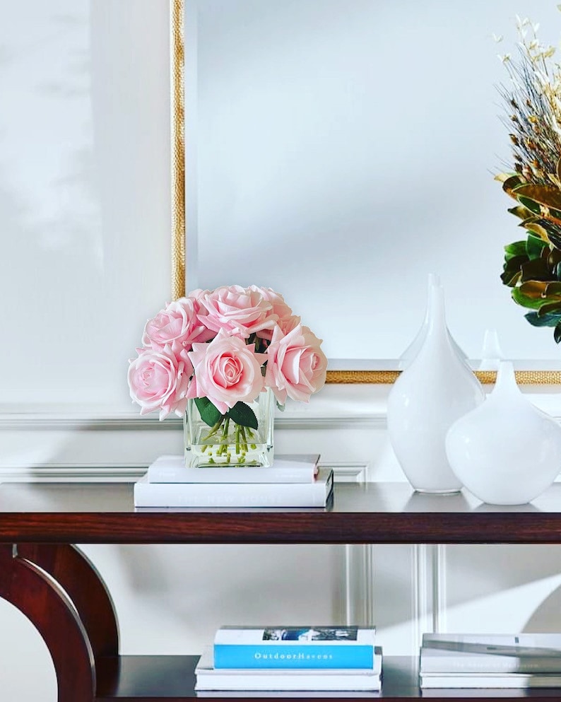May include: A wooden console table with two shelves, featuring a bouquet of pink roses in a clear glass vase. The table is decorated with books and two white ceramic vases.
