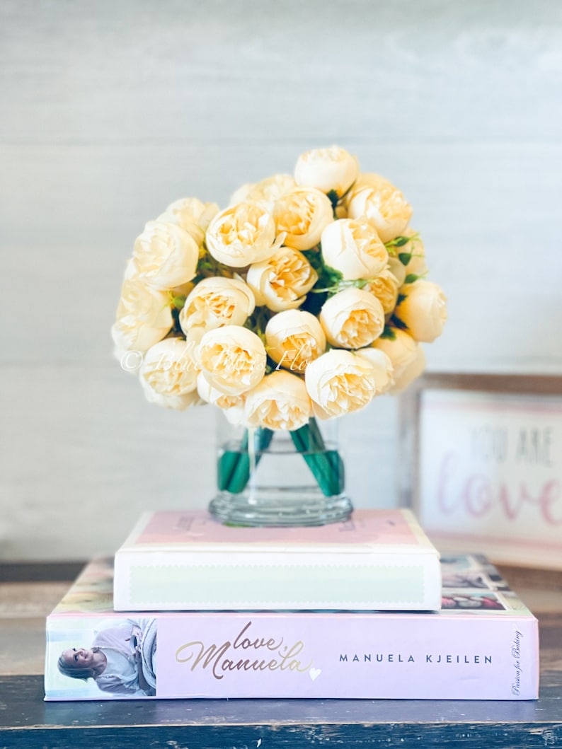 May include: A bouquet of cream-coloured artificial peonies in a clear glass vase, sitting on top of two books. The top book is white with a light green spine and the bottom book is pink with the title "love, manuela" and the author "MANUELA KJEILEN".