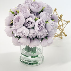 May include: A bouquet of light purple artificial peonies in a clear glass vase. The vase has a round base and is filled with water.