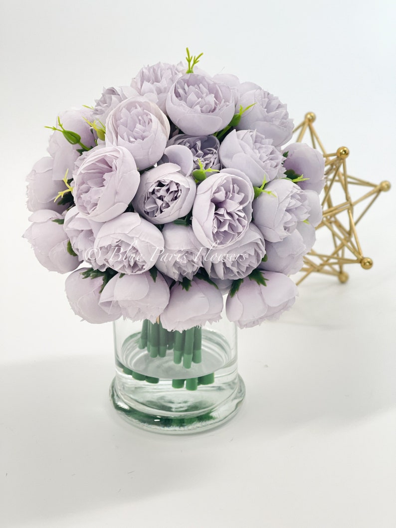 May include: A bouquet of light purple artificial peonies in a clear glass vase. The vase has a round base and is filled with water.