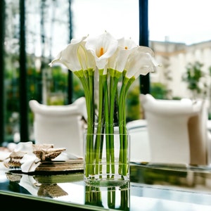 May include: A clear glass vase filled with white calla lilies with green stems. The vase is sitting on a reflective surface.