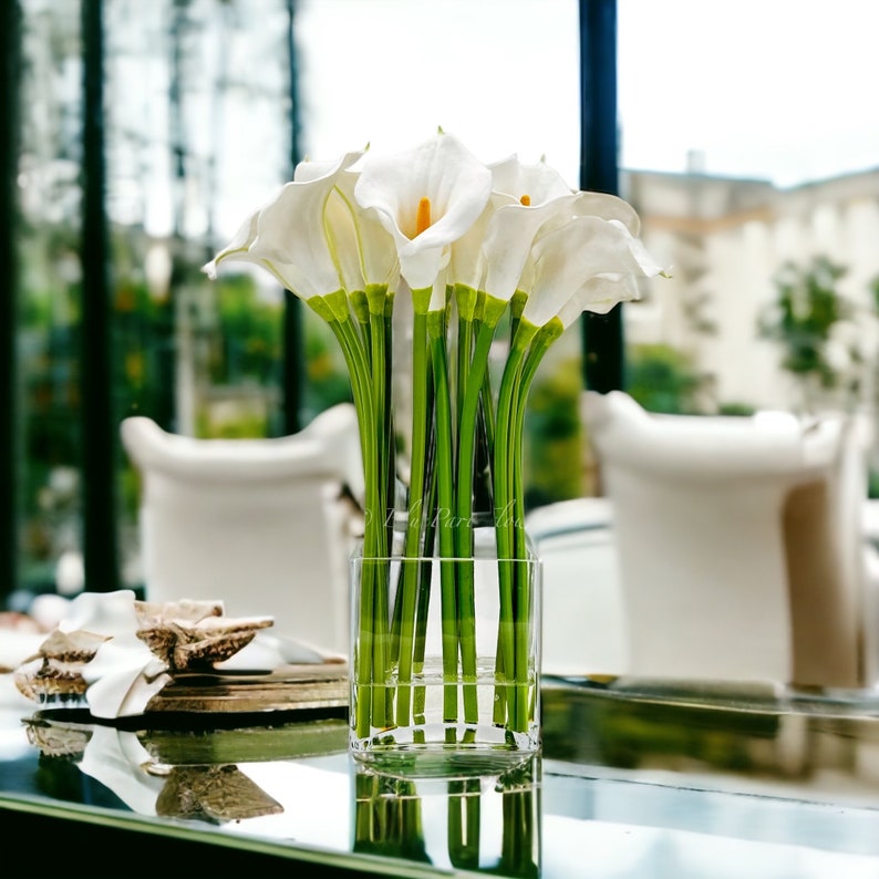 May include: A clear glass vase filled with white calla lilies with green stems. The vase is sitting on a reflective surface.