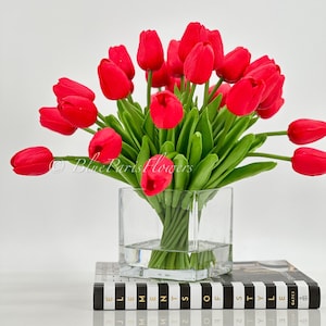 May include: A bouquet of red tulips in a clear glass vase, sitting on a black and white striped book with the text "Elements of Style" printed on the cover.