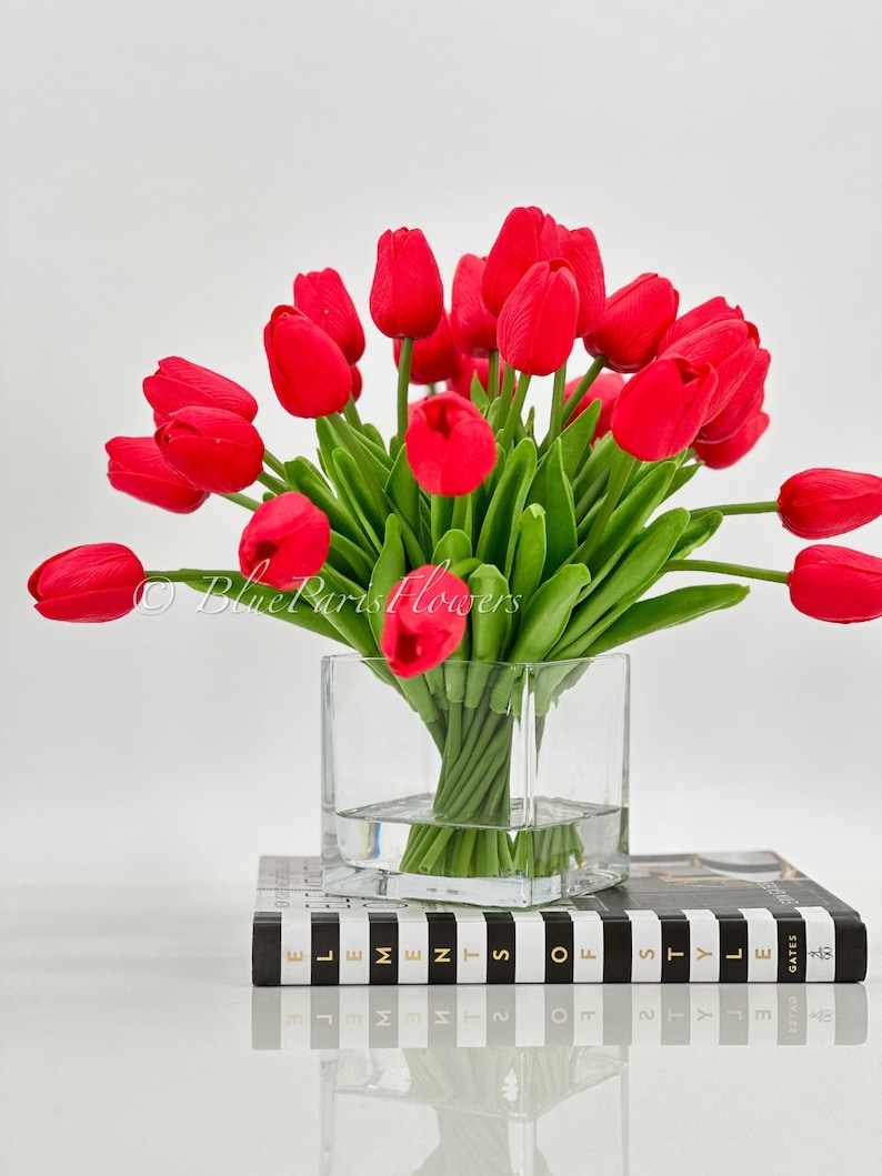 May include: A bouquet of red tulips in a clear glass vase, sitting on a black and white striped book with the text "Elements of Style" printed on the cover.