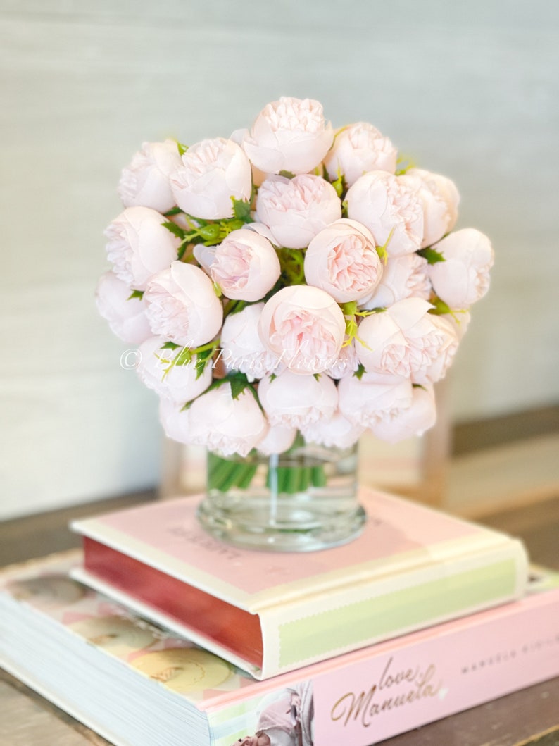 May include: A bouquet of pale pink artificial roses in a clear glass vase, sitting on top of two books. The top book is pink with the word "Bakery" printed on the cover. The bottom book is pink with the text "love,  Manuela" printed on the cover.