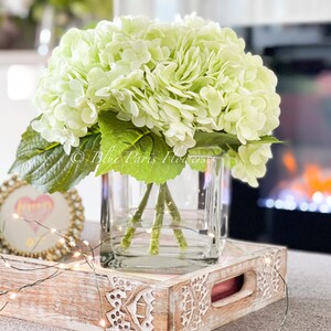 May include: A bouquet of light green hydrangeas in a clear glass vase on a white wooden tray with a string of fairy lights.