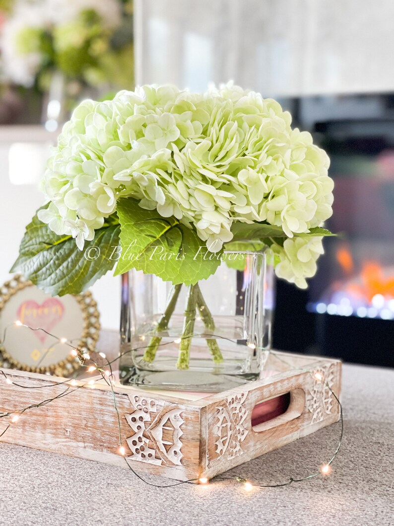 May include: A bouquet of light green hydrangeas in a clear glass vase on a white wooden tray with a string of fairy lights.