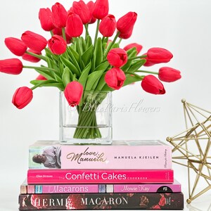 May include: A bouquet of red tulips in a clear glass vase, sitting on a stack of four books with pink and black covers. The books have titles like "Love & Manuela", "Confetti Cakes Cookbook", "Macarons", and "HERM". A gold geometric sculpture is in the background.
