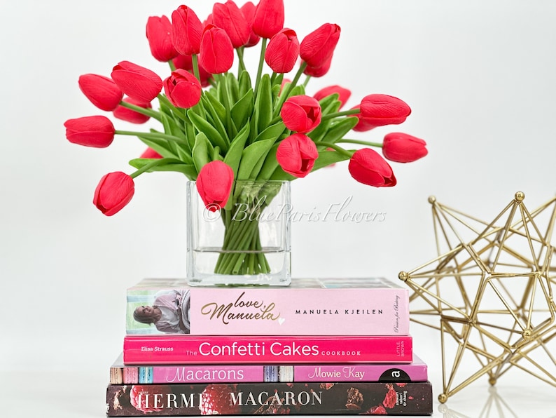 May include: A bouquet of red tulips in a clear glass vase, sitting on a stack of four books with pink and black covers. The books have titles like "Love & Manuela", "Confetti Cakes Cookbook", "Macarons", and "HERM". A gold geometric sculpture is in the background.