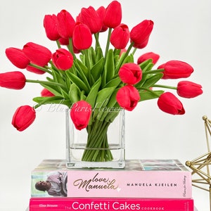 May include: A bouquet of red tulips in a clear glass vase, sitting on top of a stack of cookbooks. The books are titled "Love Manuela", "The Confetti Cakes Cookbook", "Macarons", and "Herme Macaron".