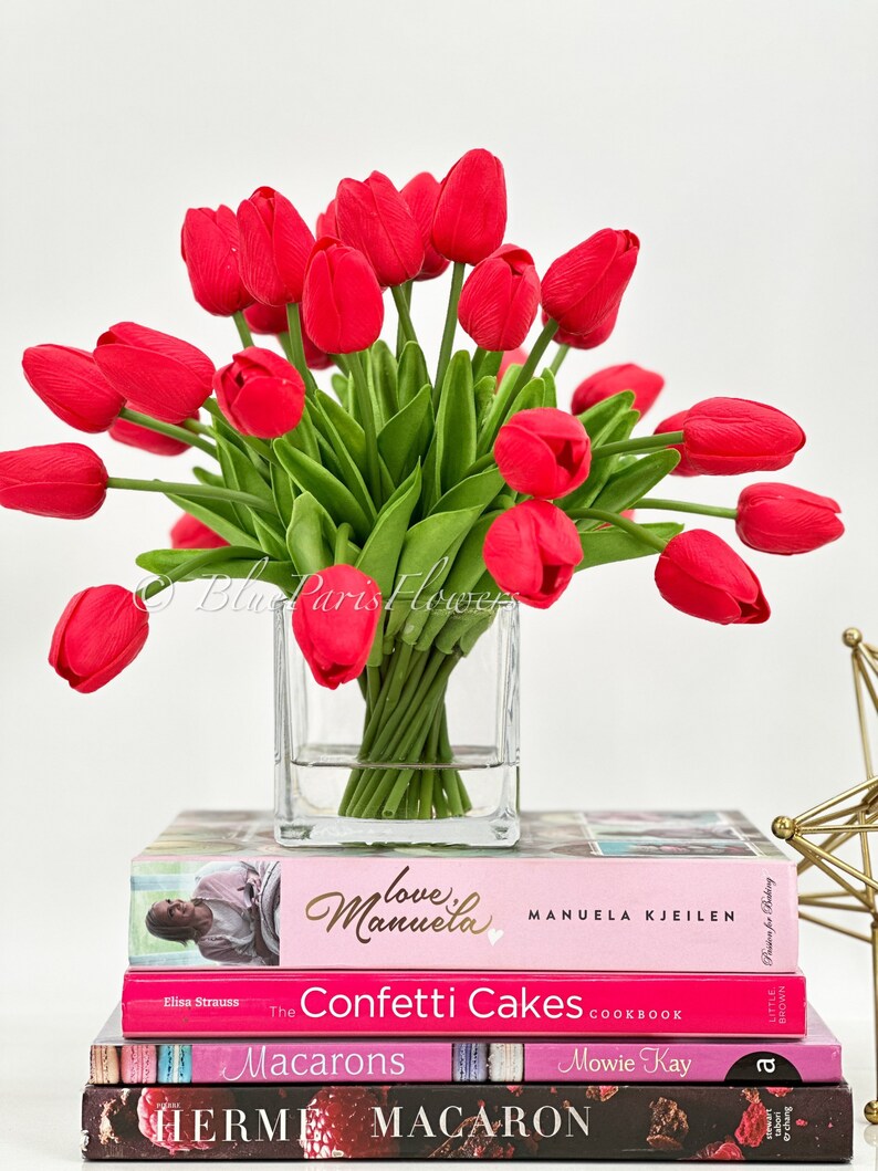May include: A bouquet of red tulips in a clear glass vase, sitting on top of a stack of cookbooks. The books are titled "Love Manuela", "The Confetti Cakes Cookbook", "Macarons", and "Herme Macaron".