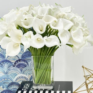 May include: A bouquet of white calla lilies arranged in a clear glass vase. The vase is sitting on a black and white striped surface.