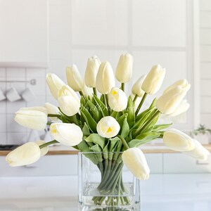 May include: A bouquet of white artificial tulips in a clear glass square vase. The tulips are arranged in a loose, natural style, with some of the stems extending beyond the vase.