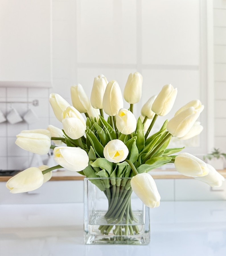 May include: A bouquet of white artificial tulips in a clear glass square vase. The tulips are arranged in a loose, natural style, with some of the stems extending beyond the vase.