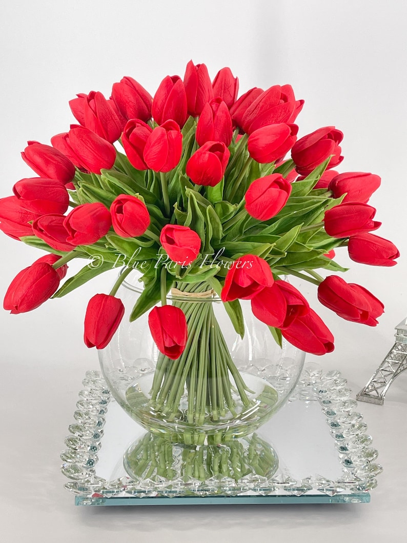 May include: A bouquet of red tulips arranged in a clear glass vase. The vase is sitting on a mirrored tray with a crystal-like border.