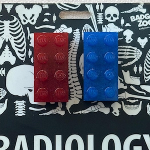 May include: A black and white Radiology badge with a skeleton and skull design, featuring the text "RADIOLOGY". Two Lego bricks, one red and one blue, are placed on top of the badge.