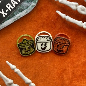 May include: Three Halloween-themed pumpkin buckets with different colored glitter. The buckets are green, white, and orange. The green bucket has a black and white face with a hat. The white bucket has a black and white face with a smile. The orange bucket has a black and white face with a frown.