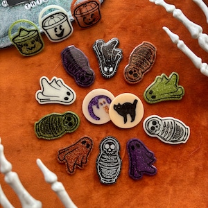 May include: A collection of 12 Halloween-themed resin pins featuring various spooky designs, including ghosts, skeletons, pumpkins, and a cat. The pins are decorated with glitter and have a variety of colors, including green, orange, purple, black, and white.