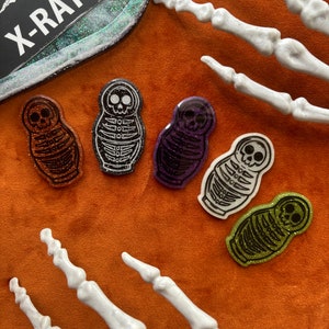 May include: A collection of colorful, glittery resin skeleton doll pins in orange, black, purple, and green. The pins are shaped like nesting dolls with a skeleton design. A decorative tray with the word "X-RAY" is in the background.