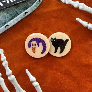 May include: Two round resin Halloween themed coasters. One coaster features a black cat silhouette and the other features a ghost with a purple and orange design.