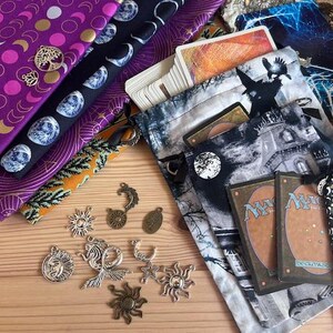 May include: Assortment of fabrics with celestial and botanical patterns, including purple, black, and orange. Silver charms with sun, moon, and tree designs are scattered around. Several card pouches and playing cards are also visible.