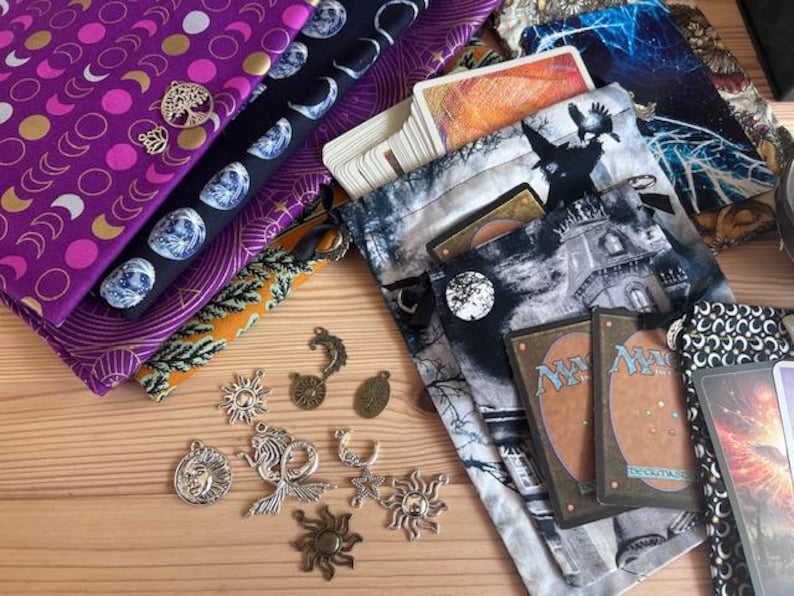 May include: Assortment of fabrics with celestial and botanical patterns, including purple, black, and orange. Silver charms with sun, moon, and tree designs are scattered around. Several card pouches and playing cards are also visible.