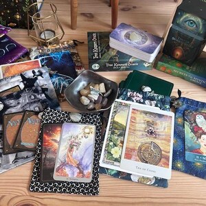May include: An assortment of tarot cards, oracle decks, and decorative pouches are arranged on a wooden surface. The cards feature colorful illustrations, and the pouches display various patterns. A small bowl of stones and a candle holder are also present.