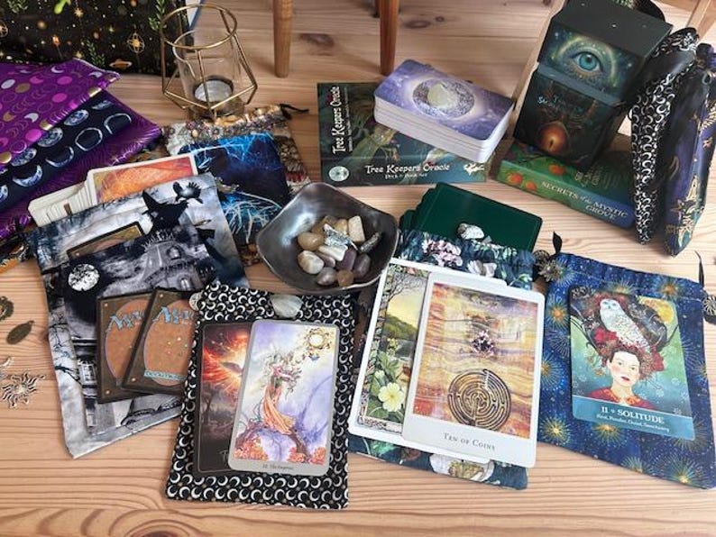 May include: An assortment of tarot cards, oracle decks, and decorative pouches are arranged on a wooden surface. The cards feature colorful illustrations, and the pouches display various patterns. A small bowl of stones and a candle holder are also present.