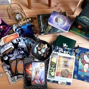 May include: An assortment of tarot cards, oracle decks, and decorative pouches are arranged on a wooden surface. The decks feature celestial and nature-themed artwork. A small bowl of stones and a decorative candle holder are also present.