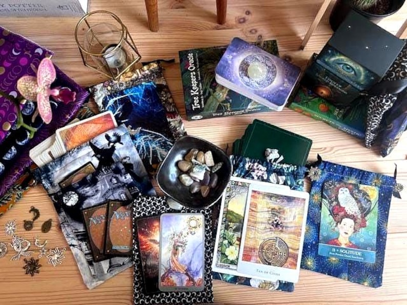 May include: An assortment of tarot cards, oracle decks, and decorative pouches are arranged on a wooden surface. The decks feature celestial and nature-themed artwork. A small bowl of stones and a decorative candle holder are also present.