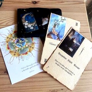 May include: A collection of tarot cards and a black velvet pouch with a celestial design. The cards feature illustrations and text, including "Deception Theft Escape" and "Notion". The artwork includes a moon, flowers, and a dreamlike mandala.