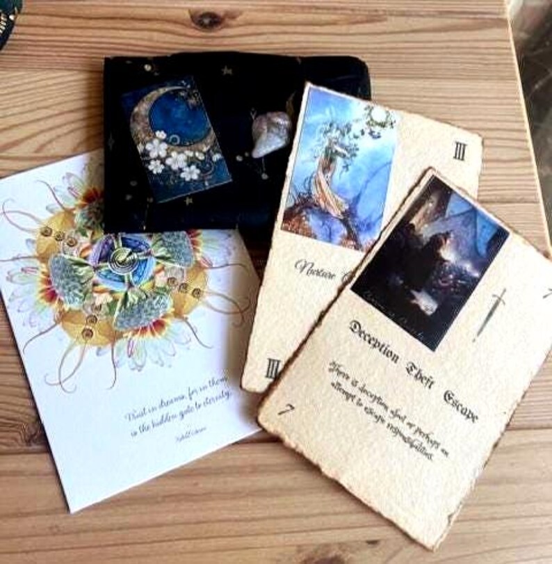 May include: A collection of tarot cards and a black velvet pouch with a celestial design. The cards feature illustrations and text, including "Deception Theft Escape" and "Notion". The artwork includes a moon, flowers, and a dreamlike mandala.