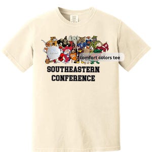 Może przedstawiać: Kremowa koszulka z grafiką różnych maskotek z kreskówek i napisem "SOUTHEASTERN CONFERENCE" czarnymi literami. Koszulka ma również napis "comfort colors tee".