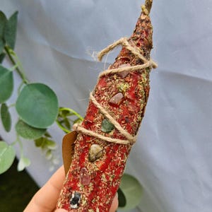 Könnte beinhalten: Ein roter Räucherstäbchen, verziert mit Kristallen und Kräutern, mit Bindfaden umwickelt. Der Stab hat eine strukturierte Oberfläche und verjüngt sich zu einem Punkt. Ein kleines Etikett ist angebracht.