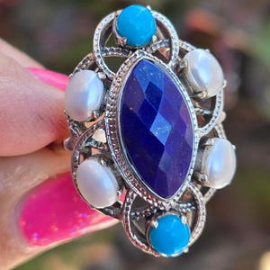 May include: Silver-toned ring featuring a large, faceted, deep blue gemstone at the center, surrounded by smaller white and turquoise stones. The ring has an ornate, filigree design. The ring is a statement piece of jewelry.