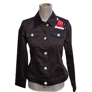 Könnte beinhalten: Schwarze Jeansjacke mit einem aufgenähten Weihnachtsmützen-Applikation auf der oberen rechten Brust. Die Jacke hat Strassknöpfe und Taschenakzente. Die Jacke wird auf einer Schaufensterpuppe präsentiert.
