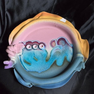 May include: A handcrafted ceramic plate with a unique design. The plate features a pink center, surrounded by a blue and turquoise pattern. The rim is a blend of yellow and orange. The plate has a textured, sculpted handle on the side.
