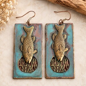 May include: A pair of rectangular drop earrings featuring a textured turquoise and copper-toned patina finish. Each earring displays a raised, gold-toned fish motif positioned above a circular cutout containing an intricate, abstract metal pattern.