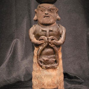 May include: A brown ceramic figurine of a bearded man with a bowl on his head. The figure has a cross on its chest and holds a loop-shaped object. The base has a small drawer. The figurine is likely a candle holder.