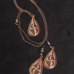 Puede incluir: Un conjunto de joyas de color cobre que incluye un collar y pendientes a juego. El colgante y los pendientes tienen forma de lágrima con un diseño geométrico recortado. El collar tiene una cadena delicada.