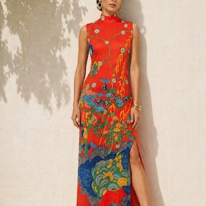 Op de afbeelding: Een mouwloze, rode maxi-jurk met een hoge halslijn en een split tot aan de dij. De jurk heeft een kleurrijk bloemen- en landschapspatroon in blauw, geel en groen. Gouden accessoires maken de look compleet.