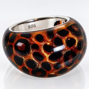 May include: A wide, rounded ring featuring a glossy, amber-colored resin exterior with a black leopard print pattern and gold-toned glitter accents. The interior of the band is smooth, polished silver metal with the hallmark 925 stamped inside.
